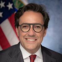 A professional headshot of a man wearing glasses, a dark suit, white shirt, and red tie. He is smiling slightly and posed in front of a blurred background featuring an American flag.